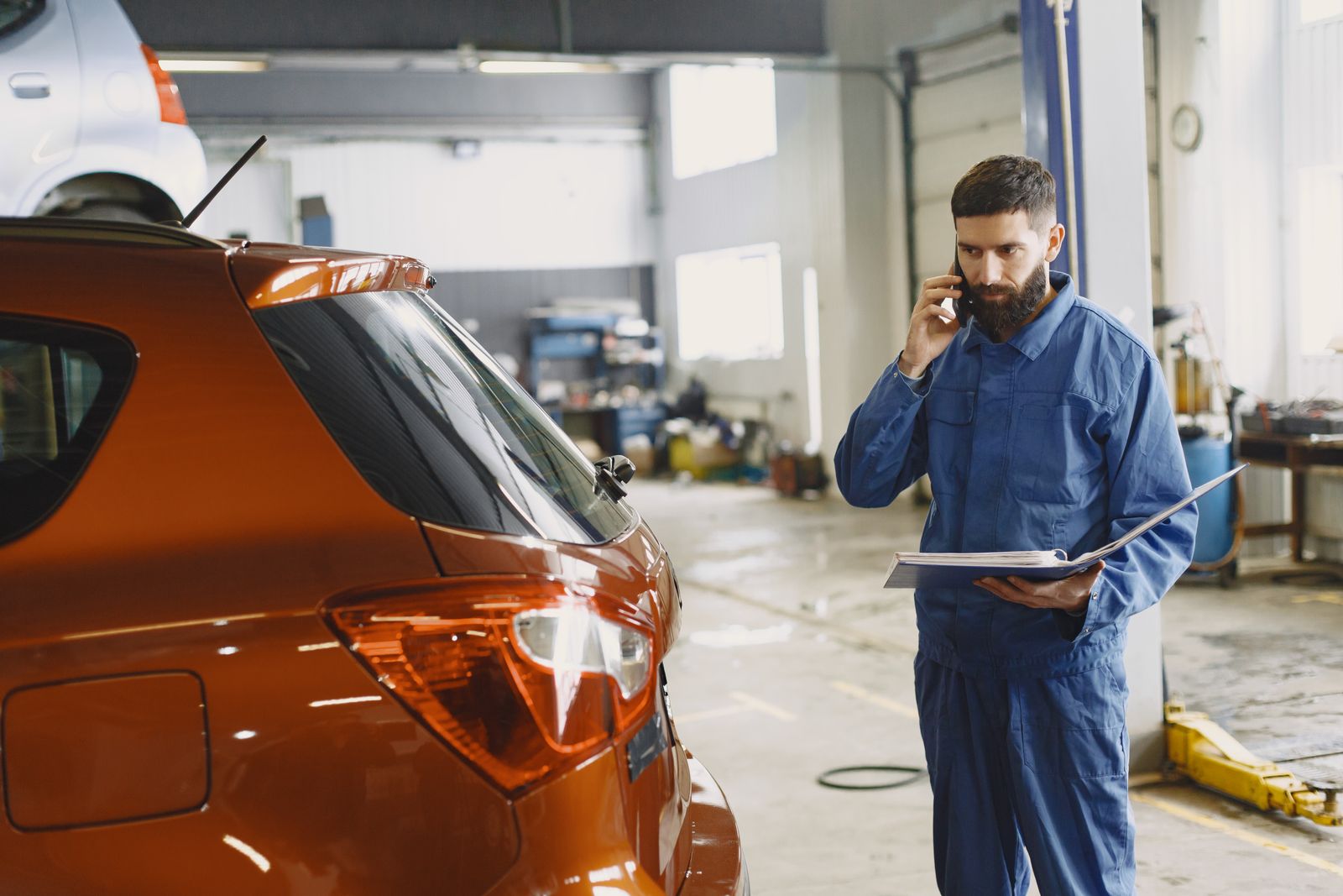 Handwerker telefoniert mitten in der Arbeit in seiner Werkstatt, Auftragsmappe in der Hand